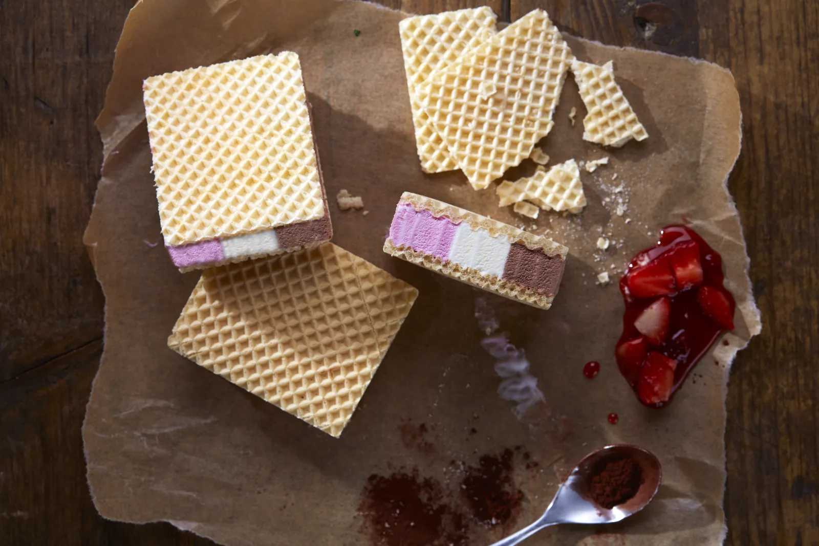 Eiswaffel-Sandwich mit Erdbeer-Vanille-Schoko-Streifen neben Waffelbruch und Erdbeeren